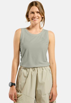 Femme souriante portant un haut de sport sans manches gris clair et un pantalon cargo beige, avec une smartwatch noire à son poignet gauche.