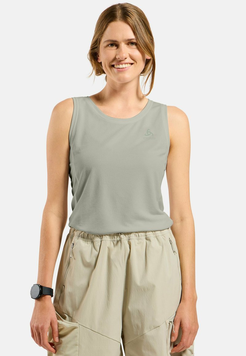 Lächelnde Frau, die ein hellgraues ärmelloses Sporttop und beige Cargohosen trägt, mit einer schwarzen Smartwatch am linken Handgelenk.