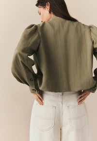 Oliivroheline lühikesed varrukad ja nööpide aktsentidega crop blouse, kombineeritud kõrge vöökohaga valgete teksadega, millel on tagataskud ja kollased õmblused.