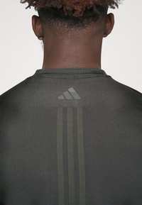 Camisa atlética gris con cuello redondo, tres franjas verticales en verde y un logo de Adidas en un tono más oscuro en la parte posterior.