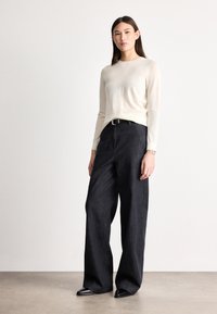 Top bej deschis cu mâneci lungi, asortat cu pantaloni evazați din denim închis, cu talie înaltă. Pantofi negri cu vârf ascuțit; design minimalist cu textură subtilă.