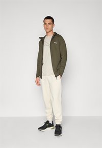 Sweat à capuche zippé olive, t-shirt gris clair avec un motif en lignes, jogging crème et baskets noires avec des accents verts. Tenue de sport simple.