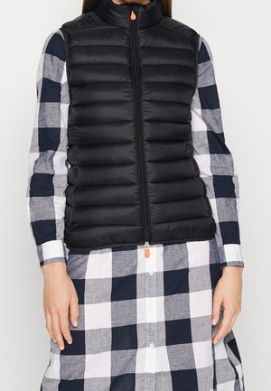 Personne portant une veste noire matelassée par-dessus une robe chemise à manches longues à carreaux noirs et blancs, debout devant un fond uni.