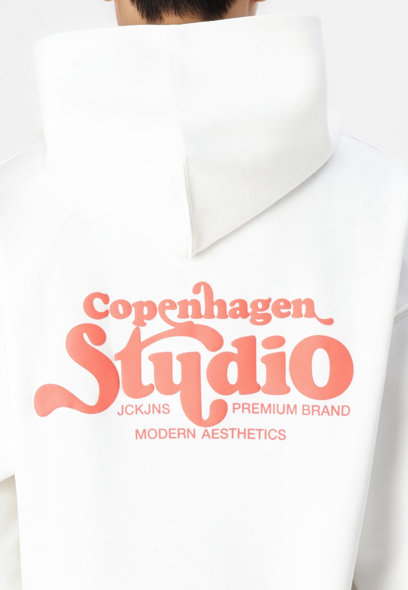 Witte hoodie met een grote roze graphic met de tekst "Copenhagen Studio" en aanvullende branding eronder. Het materiaal lijkt zacht en heeft een relaxte fit.
