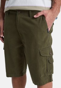 Man in olijfgroene cargoshorts met meerdere zakken, met één hand in de voorzak, gecombineerd met een beige gestreept shirt.