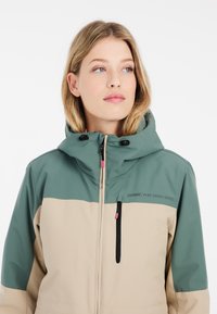 Zweifarbige Outdoor-Jacke mit grünem Kapuze und Oben, beigem Unterteil, mit Reißverschlusstaschen und verstellbaren Verschlüssen.