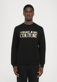 Črna majica s dolgimi rokavi in okroglim vratom. Na sprednji strani je zlati napis "VERSACE JEANS COUTURE". Mehka tkanina, zasnova sproščenega kroja.