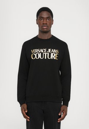 Black sweatshirt with long sleeves and crew neck. Features gold text "VERSACE JEANS COUTURE" on the front. Soft fabric, relaxed fit design.