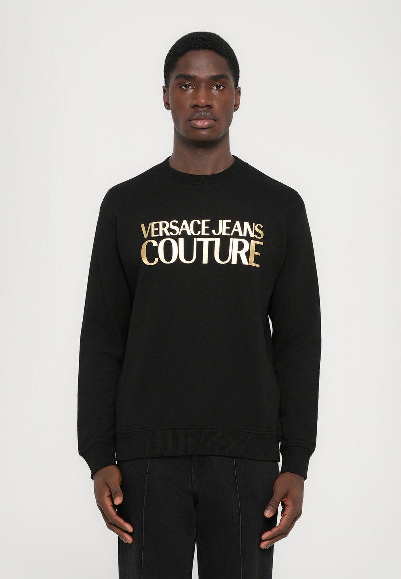 Črna majica s dolgimi rokavi in okroglim vratom. Na sprednji strani je zlati napis "VERSACE JEANS COUTURE". Mehka tkanina, zasnova sproščenega kroja.