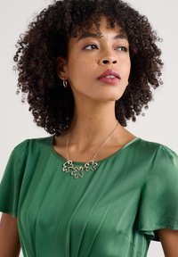 Grünes Kleid mit kurzen Ärmeln, glattem Stoff und rundem Ausschnitt. Besitzt eine goldene Halskette mit blattförmigen Akzenten.
