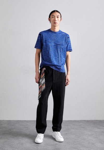 T-shirt azul listrado com mangas curtas combinado com calças pretas texturizadas, apresentando um lenço estampado colorido como destaque na cintura. Ténis brancos.