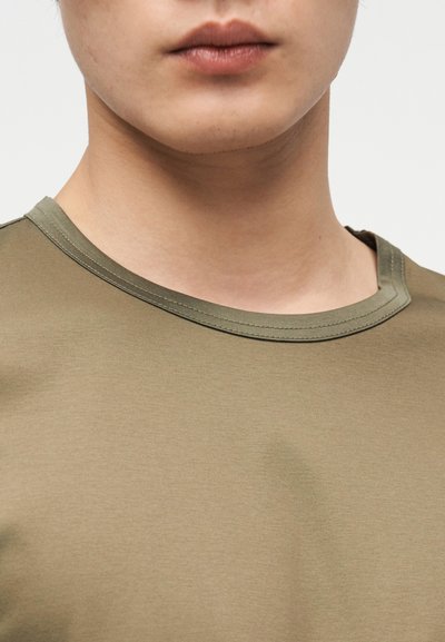 Κοντινό πλάνο του κάτω μέρους του προσώπου και του λαιμού ενός ατόμου που φοράει πουκάμισο ελιάς με στρογγυλή λαιμόκοψη, σε απλό φωτεινό φόντο.