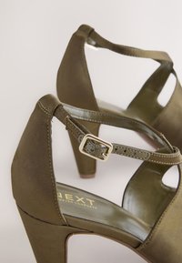 Oliivirohelised kõrgete kontsadega sandaalid, millel on sile tekstuur, pahkluu rihm ja kuldse pandlaga kaunistus. Siseküljel on kuldse logoga silt.