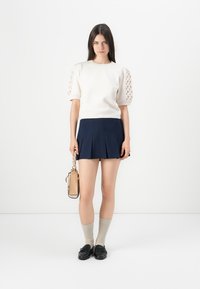 Witte korte mouwtrui met gestructureerde pofmouwen, gecombineerd met een marineblauwe geplooide rok. Model houdt een kleine tan tas vast en draagt kniehoge sokken.