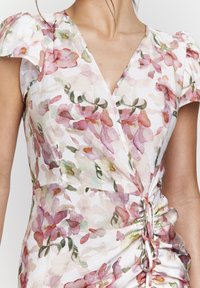 Robe portefeuille florale en tissu léger, arborant des teintes de rose et de vert, avec de courtes manches volantées et un détail froncé sur le côté avec cordon de serrage.