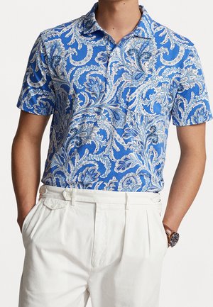 Homme portant un polo bleu à manches courtes avec un motif cachemire blanc et un pantalon blanc plissé, les mains dans les poches.