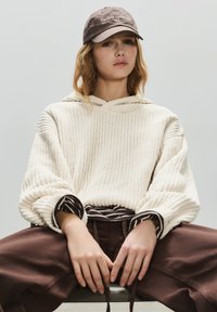 Hanorac tricotat crem cu mâneci largi, textură ușoară; purtat peste o cămașă maro cu dungi. Șapcă maro închis cu detaliu text. Pantaloni maro.