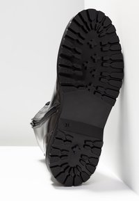 Suela de bota negra con profundos surcos y patrones en forma de cruz, talla 37, inclinada contra una superficie blanca.