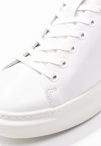 Białe skórzane trampki z płaskimi białymi sznurówkami, okrągłym noskiem, minimalistycznym designem i teksturowaną gumową podeszwą. Subtelne detale haftu po bokach.