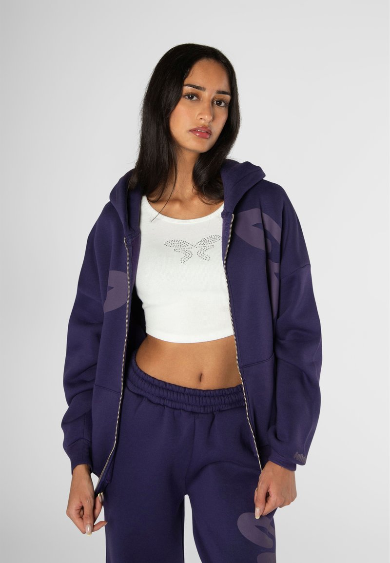 Felpa viola con zip e grandi dettagli del logo, abbinata a un crop top bianco con motivo farfalla e pantaloni jogger viola coordinati.