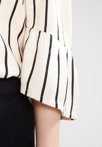 Krémová blůza s černými vertikálními pruhy, s rozšířenými rukávy a texturovanou tkaninou. Design zahrnuje upravené detaily na lemech.
