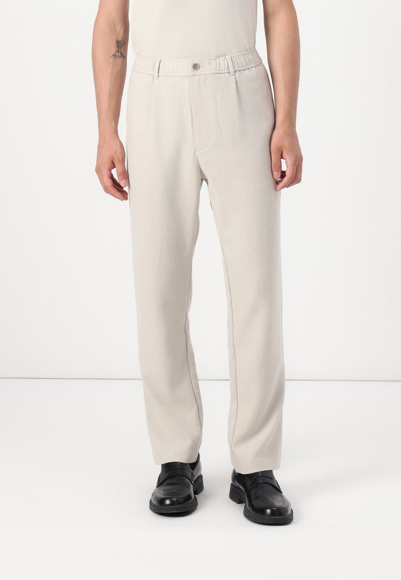 Light beige, straight-leg trousers with a soft, textured fabric. Features an elastic waistband and a single button closure. Worn with black shoes.