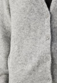 Cardigan grigio realizzato in tessuto a maglia testurizzato, con chiusura a bottoni e quattro bottoni in plastica grigia chiara, e una tasca sul lato sinistro.