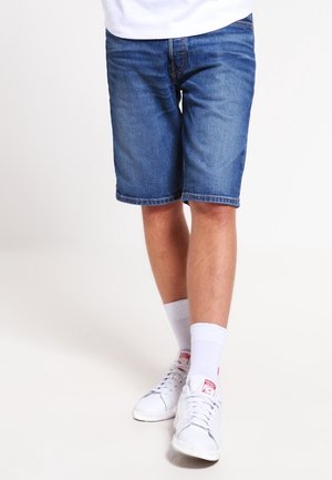 Osoba w niebieskich dżinsowych szortach, białych skarpetkach i białych trampkach, stojąca z jedną nogą lekko wysuniętą do przodu na białym tle.