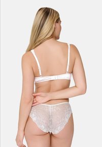 Donna con lunghi capelli biondi, che indossa un reggiseno di pizzo bianco e mutandine di pizzo abbinate, fotografata di spalle su sfondo grigio chiaro.