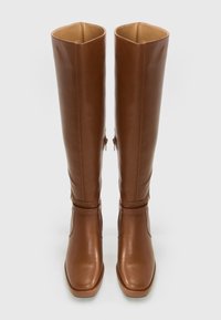 Botas de cuero marrón hasta la rodilla con punteras cuadradas, cremallera en los lados internos y una correa decorativa alrededor del tobillo.