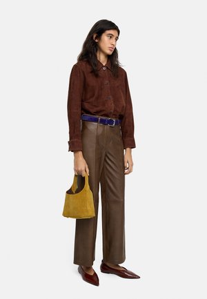 Osoba ubrana w brązową zamszową koszulę, brązowe skórzane spodnie z wysokim stanem i niebieskim paskiem, trzymająca musztardową zamszową torebkę oraz nosząca brązowe pointed buty, na białym tle.