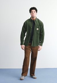 Zelena korduroy košulja s lijevim džepom, nosi se preko crne košulje, uparen s smešnim hlačama. Outfit je upotpunjen sivoj tenisicama.