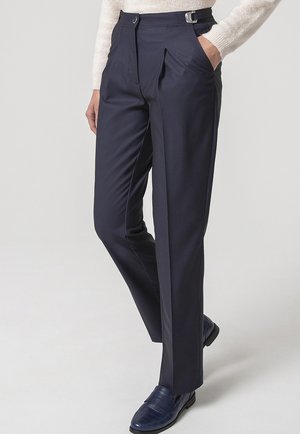 Persona che indossa pantaloni blu navy su misura con dettaglio fibbia laterale, maglione bianco e mocassini blu scuro, in piedi con una mano in tasca.