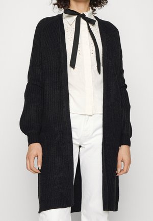 Person trägt eine lange schwarze Strickjacke über einer weißen Bluse mit schwarzer Schleife und weißen Hosen, steht vor einem einfarbigen Hintergrund.