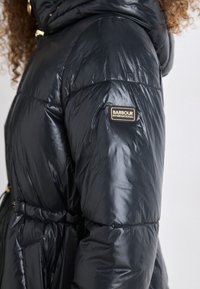Casaco puffer preto com textura brilhante, cintura ajustada e um patch de logótipo na manga. Tem capuz e detalhes em hardware dourado.