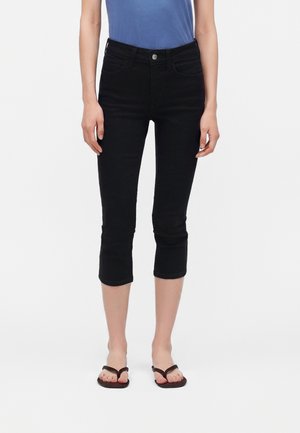 Kvinde iført sorte korte skinny jeans, blå top og mørkebrune flipflops står foran en ensfarvet hvid baggrund.