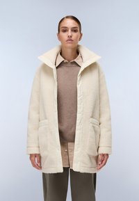 Casaco exterior de cor creme com uma textura peluda, colarinho oversized, dois bolsos frontais e fecho de botão, sobre um suéter castanho.