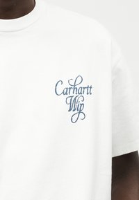 Λευκό T-shirt με στρογγυλή λαιμόκοψη που φοριέται από άτομο, με μικρό μπλε στυλιζαρισμένο κείμενο "Carhartt Wip" στο αριστερό στήθος.