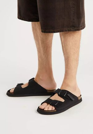 Piernas que llevan pantalones cortos marrones y sandalias negras abiertas con dos correas ajustables con hebilla, de pie sobre una superficie clara.