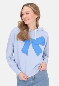 Világoskék kapucnis pulóver, amelyen egy nagy kék szalag grafika díszeleg, puha anyagból készült, laza szabású és húzózsinórral ellátott kapucnival.
