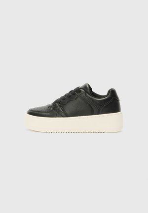 Zwarte leren sneaker met dikke witte platforms zool en zwarte veters, van opzij getoond op een effen lichtgrijze achtergrond.