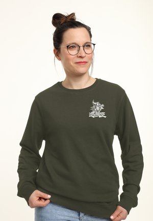 Kvinde med briller og hår i en knold bærer en mørkegrøn sweatshirt med lille hvid grafik og tekst på venstre bryst, stående mod en ensfarvet baggrund.