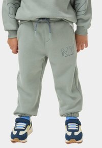 Pantalones de chándal verdes de forro polar con cintura elástica, bolsillos laterales y detalle bordado de "NY". Combinados con zapatillas azul y blanca.