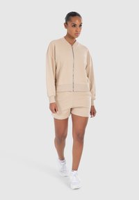 Sudadera beige con cremallera y hombros caídos, combinada con pantalones cortos a juego. El material parece suave, con un sutil detalle de logo en ambas piezas.