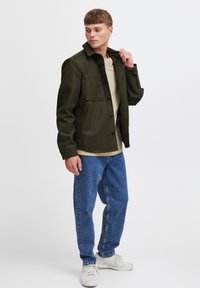 Giacca verde oliva con collo e chiusura a bottoni, due tasche sul petto, abbinata a una camicia beige e jeans in denim blu.