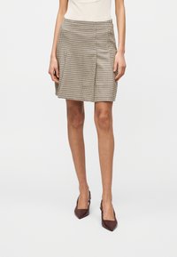 Γυναίκα που φοράει μπεζ και καφέ φούστα μέχρι το γόνατο με μοτίβο κουκίδας και φιόγκου, λευκό τοπ και καφέ τακο ύνια με ακρωτηριασμό στη φτέρνα, στέκεται μπροστά σε λευκό φόντο.