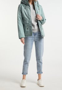 Persoon in een lichtblauwe hoodie-jack over een grijze sweatshirt, met opgerolde lichtblauwe jeans en witte sneakers, staande tegen een effen achtergrond.