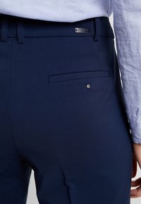 Marinblå skräddarsydda byxor med en elegant design, som har en bakficka och en diskret metallaccent på midjebandet. Slät tygtextur.