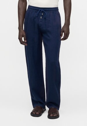 Homme portant un pantalon à cordon bleu marine et des sandales marron, debout devant un fond clair uni.