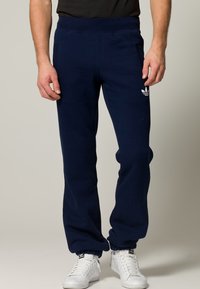 Mörkblåa sweatpants med mjuk textur, utrustade med elastisk midja och muddar, samt en vit Adidas-logga på vänstra låret.